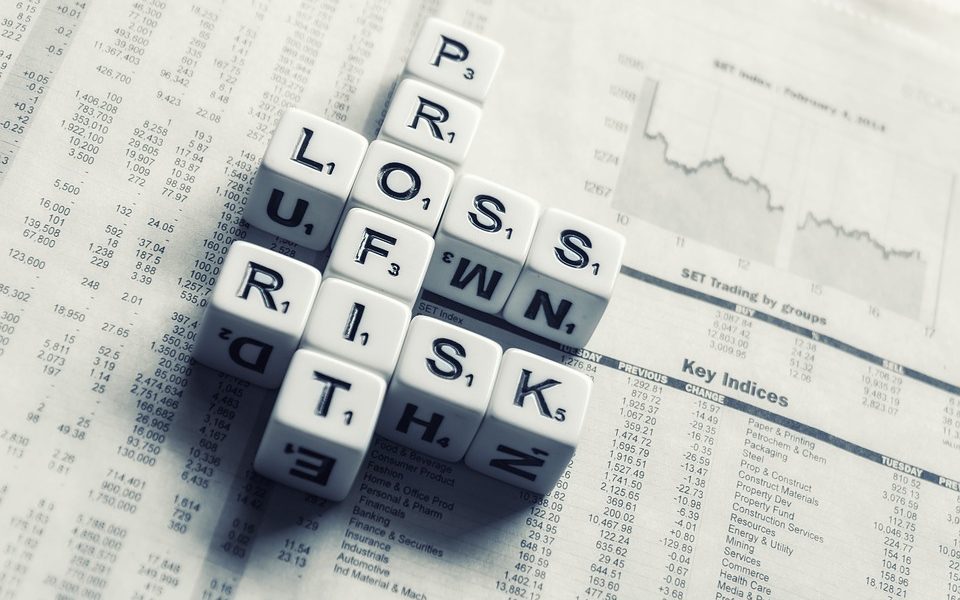 Doze dados com letras ao invés de números posicionados em cima de um jornal de finanças. Os dados formam as palavras "profit", "loss" e "risk", que em português significam lucro, perda e risco, respectivamente.