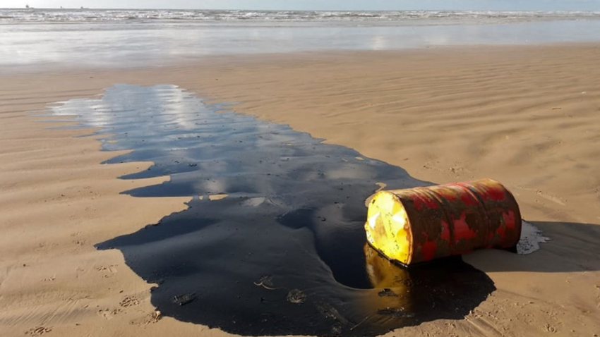 Um barril com petróleo derramado na areia da praia com o mar ao fundo.