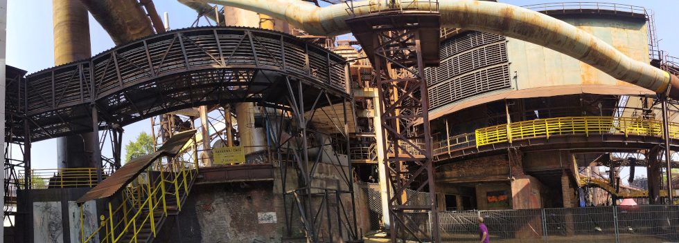 Indústria de ferro de Ostrava, com grandes tubulações enferrujadas e escadas e passarelas com corrimões amarelos.
