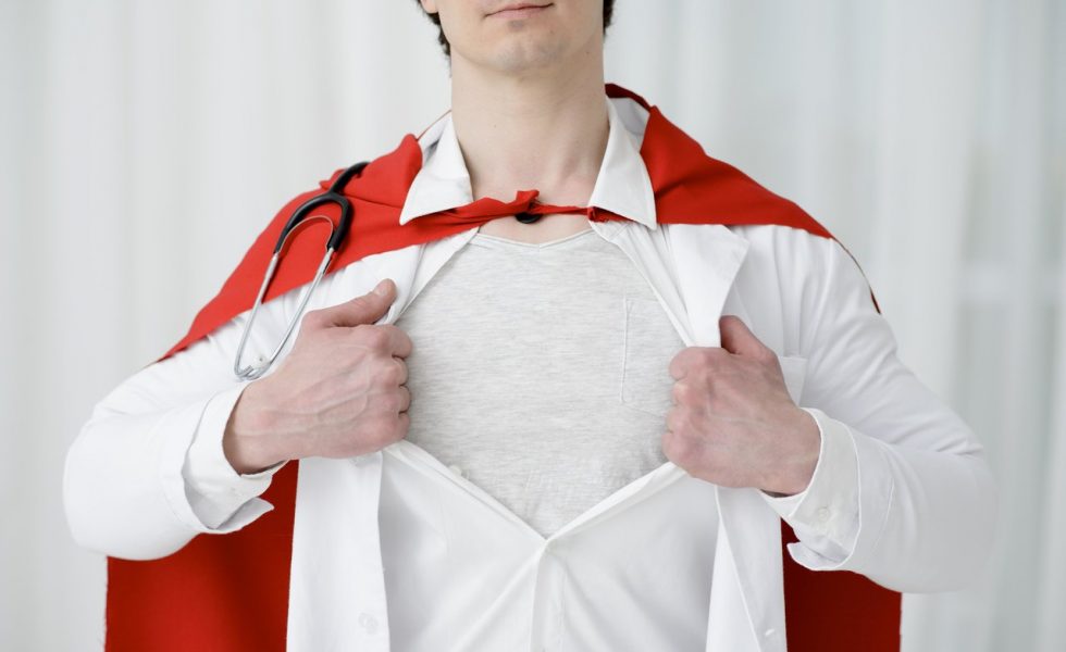 Foto de um médico da metade do rosto até a barriga. Ele é branco e está abrindo a camisa que está por baixo do jaleco e mostrando uma camiseta branca, imitando a forma como o Superman abre a camisa e mostra o uniforme. O médico ainda veste uma capa vermelha amarrada ao pescoço e tem um estetoscópio pendurado.