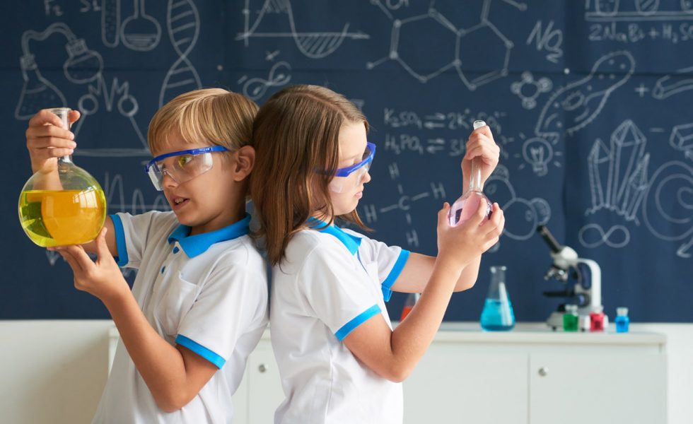 Duas crianças, um menino e uma menina, em frente a uma lousa com fórmulas e desenhos relacionados a ciência. Cada um segura um recipiente de vidro com líquido dentro.