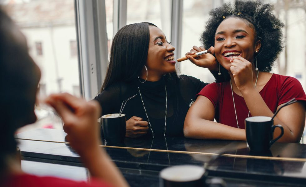 Duas mulheres negras em uma cafeteria se olhando no espelho e sorrindo enquanto uma delas maquia a outra.