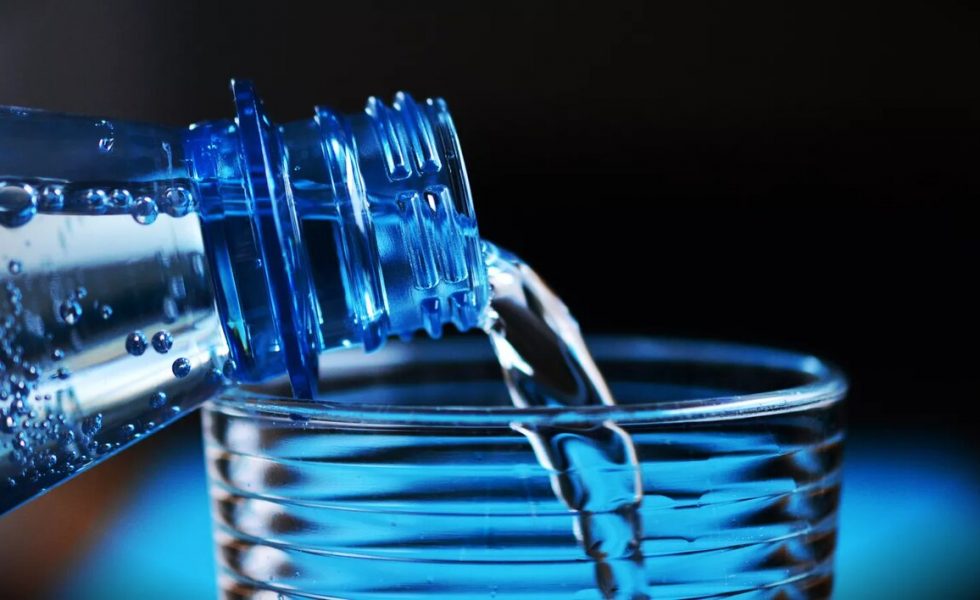 Imagem com fundo preto e detalhe azul claro na parte de baixo, uma garrafa plástica azul despejando água em copo de vidro transparente.