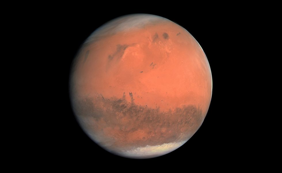 Imagem do planeta Marte, com cor avermelhada, em um fundo preto. Seus polos aparecem esbranquiçados devido à presença de calotas de gelo.