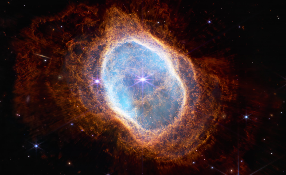Estrela brilhante central, da cor roxo-azul, rodeada de um halo iridescente branco-azulado, o qual termina numa forma de anel deformado, a partir do qual é possível observar uma espécie de pó laranja-avermelhado, tudo isto contra um fundo preto povoado de estrelas.