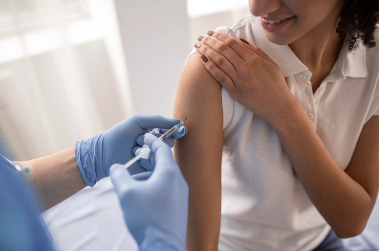 À direita uma mulher, jovem, branca de cabelos enrolados e vestindo blusa branca e levanta a manga para tomar uma vacina, sendo aplicada por uma pessoa usando uma luva azul.