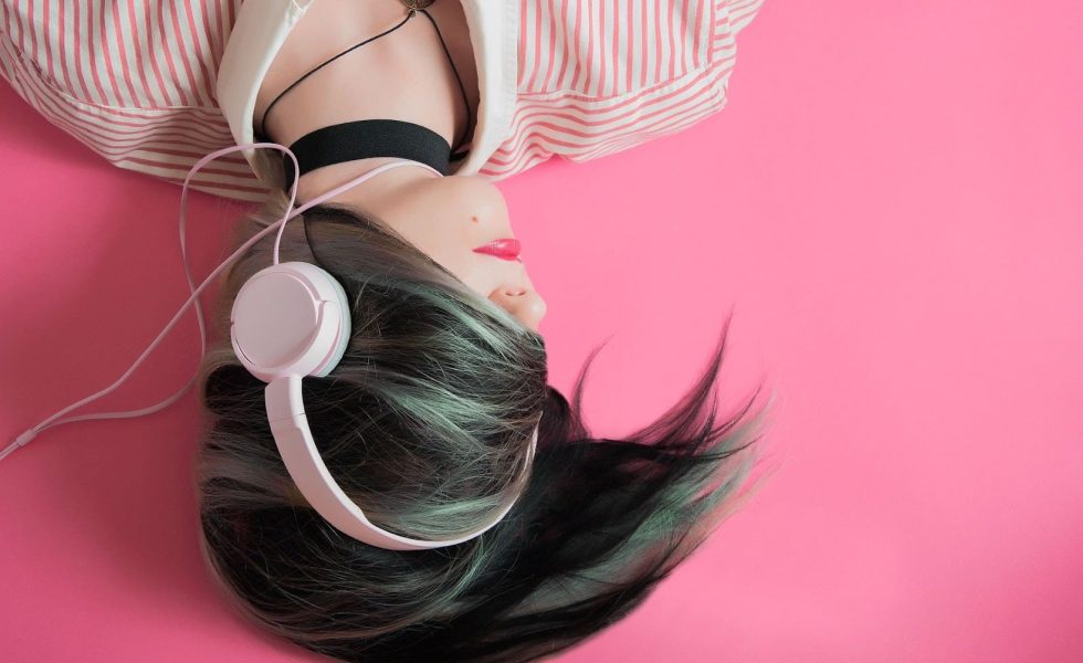 Imagem de menina, de ponta cabeça, deitada em um fundo rosa, ouvindo música por um headphone de forma que o cabelo dela cubra seus olhos.
