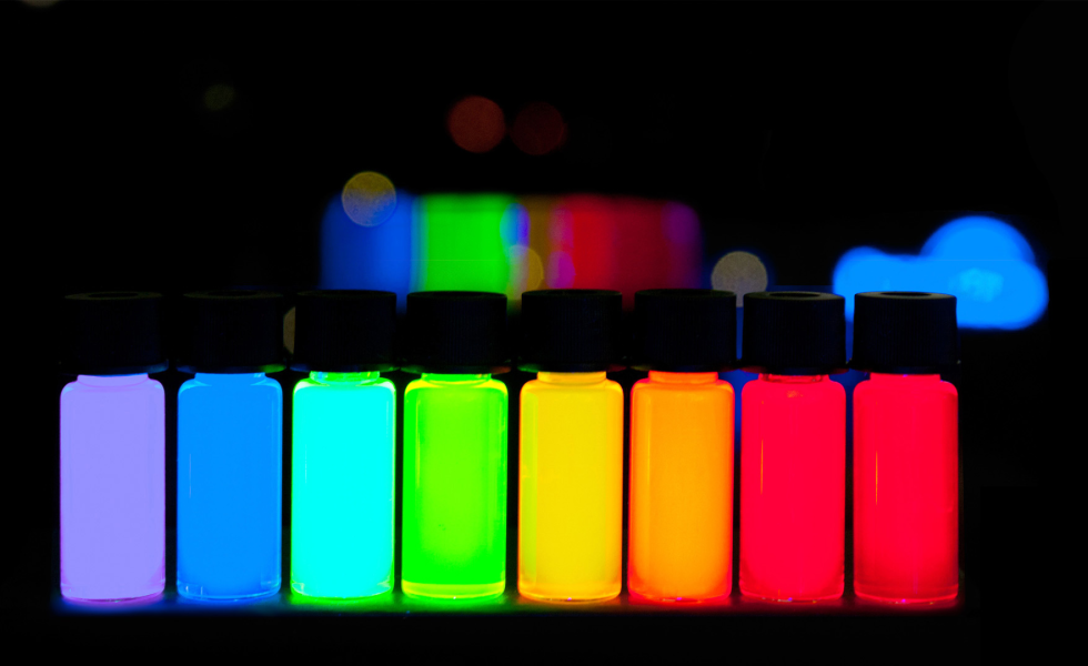 Imagem de fundo preto onde se observam uma série de frasquinhos contendo soluções com cores intensas e brilhantes, na ordem: violeta, azul, turquesa, verde, amarelo, laranja e vermelho.