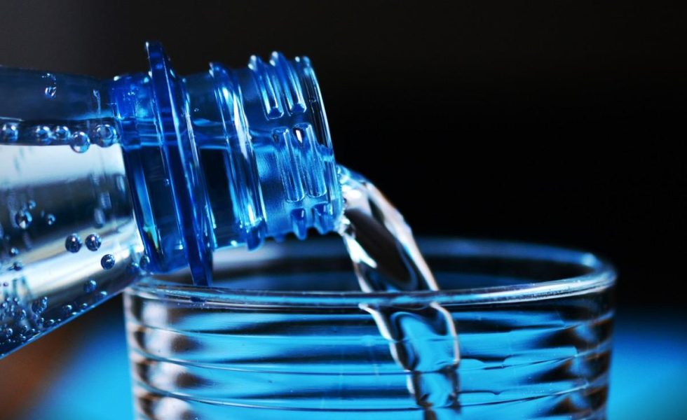 Foto em alta resolução de tons azulados e foco fechado na boca de um copo de vidro recebendo água de uma garrafa plástica.